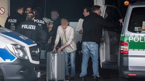 Mit Handschellen gefesselt schiebt ein Mann am Flughafen Frankfurt seinen Rollkoffer zwischen Polizisten hindurch, während ein anderer seine Habseligkeiten in Pappkartons auf dem Arm trägt.