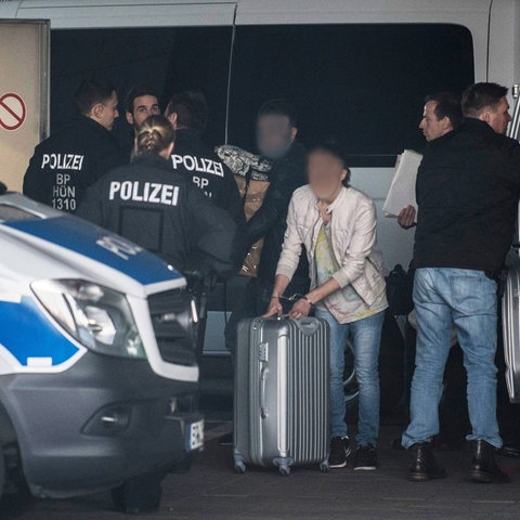 Mit Handschellen gefesselt schiebt ein Mann am Flughafen Frankfurt seinen Rollkoffer zwischen Polizisten hindurch, während ein anderer seine Habseligkeiten in Pappkartons auf dem Arm trägt.