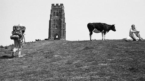 Martin Parr: Glastonbury Tor, Somerset, England, 1975 - Auf einem Hügel steht ein Turm, daneben eine Kuh und zwei Menschen (Schwarzweißbild)